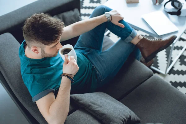 Blick aus der Vogelperspektive auf einen jungen Mann, der Kaffee trinkt, während er auf der Couch sitzt — Stockfoto