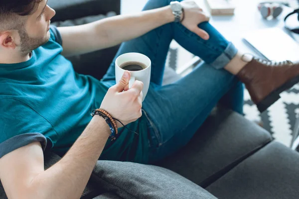 Обрезанный снимок стильного молодого человека с чашкой кофе и сидящего на диване — стоковое фото