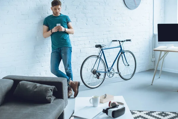 Schöner junger Mann lehnt an Wand und benutzt Smartphone zu Hause — Stockfoto
