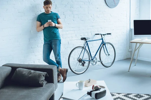 Jovem bonito inclinando-se na parede e usando smartphone em casa — Fotografia de Stock