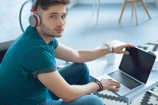 Jovem freelancer bonito em fones de ouvido usando laptop e olhando para a câmera em casa — Fotografia de Stock