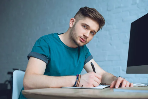Joven guapo tomando notas y mirando a la cámara mientras trabaja en casa - foto de stock