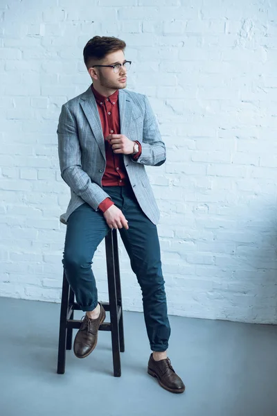 Attractive bearded man wearing suit and glasses and sitting on chair in front of white wall — Stock Photo