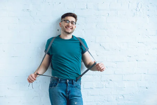 Homme barbu souriant portant un jean avec des bretelles devant un mur de briques — Photo de stock