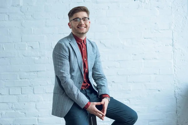 Homme élégant et souriant en vêtements décontractés assis devant un mur de briques — Photo de stock