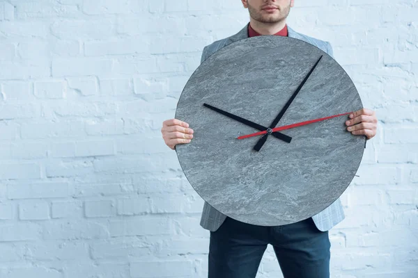 Joven sosteniendo un gran reloj delante de él mientras está de pie junto a la pared de ladrillo - foto de stock