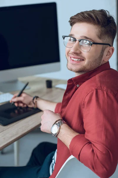 Молодой улыбающийся человек за рабочим столом с графическим планшетом и компьютером — стоковое фото