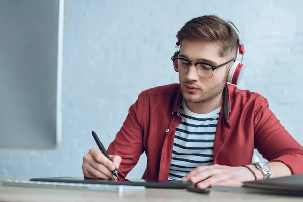 Freelancer man in headphones drawing with graphic tablet by table with computer — Stock Photo