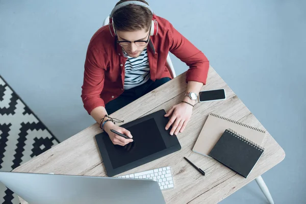 Jeune homme dans les écouteurs en utilisant tablette graphique par table avec ordinateur — Photo de stock