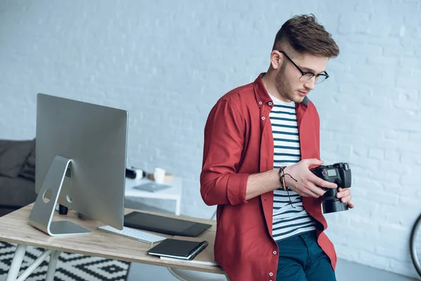 Молодой человек держит камеру и опирается на стол с компьютером в домашнем офисе — стоковое фото