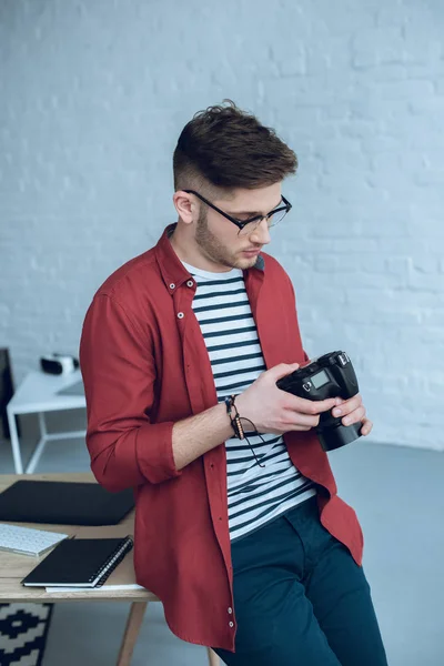 Joven barbudo en gafas sujetando cámara por mesa de trabajo - foto de stock