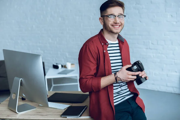 Happy freelancer holding camera by working table with computer — Stock Photo