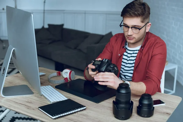 Hombre freelancer con cámara en las manos en el trabajo por mesa con ordenador y tableta gráfica - foto de stock