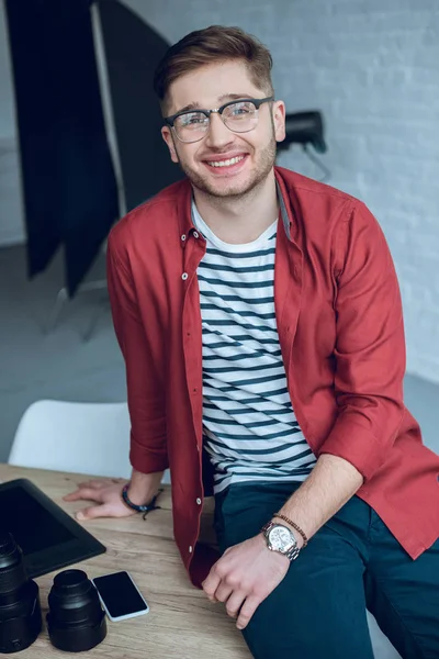 Jovem sorridente apoiado na mesa com lentes de câmera — Fotografia de Stock