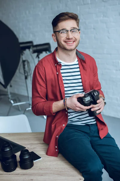 Smiling man holding camera and sitting on table with computer at home office — Stock Photo