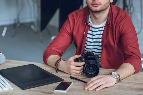 Joven barbudo sosteniendo cámara por mesa con computadora - foto de stock