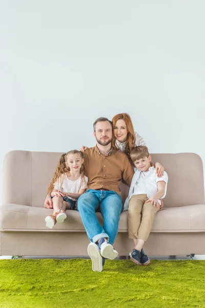 Красивая счастливая семья с двумя детьми, сидящими на диване и улыбающимися перед камерой на сером — стоковое фото