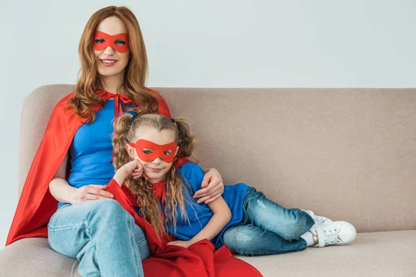 Mère heureuse et fille en costumes de super héros assis sur le canapé et souriant à la caméra — Photo de stock