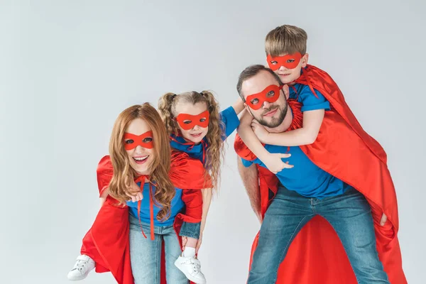 Super parents piggybacking kids pretending to be superheroes isolated on grey — Stock Photo