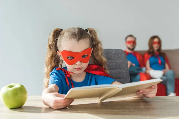 Girl in superhero costume reading book while super parents sitting behind — Stock Photo