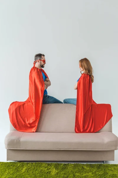 Super couple in masks and cloaks sitting on couch and looking at each other on grey — Stock Photo
