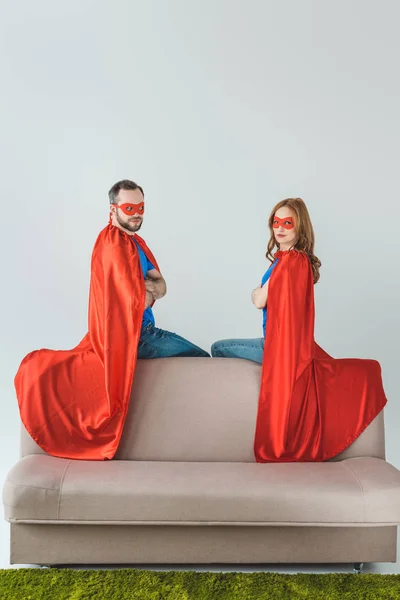 Super couple in masks and cloaks sitting on sofa and looking at camera — Stock Photo