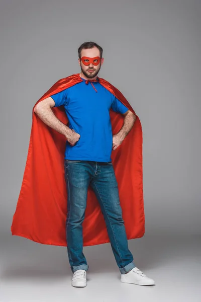 Homme en manteau rouge et masque debout avec les mains à la taille et regardant la caméra sur gris — Photo de stock