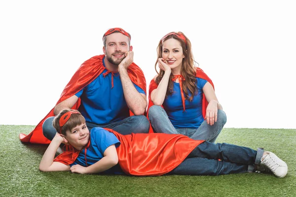 Família feliz de super-heróis sentados no gramado e sorrindo para a câmera no branco — Fotografia de Stock