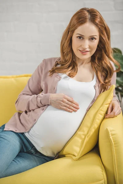 Hermosa mujer embarazada tocando el vientre y sonriendo a la cámara en casa - foto de stock