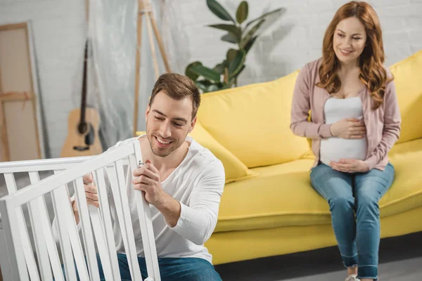 Sorrindo mulher grávida sentada no sofá e olhando para o marido feliz consertando cama de bebê — Fotografia de Stock
