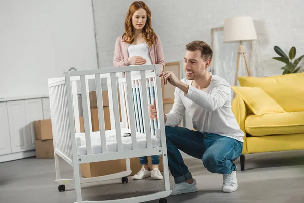 Pregnant woman looking at smiling husband fixing baby bed in new apartment — Stock Photo
