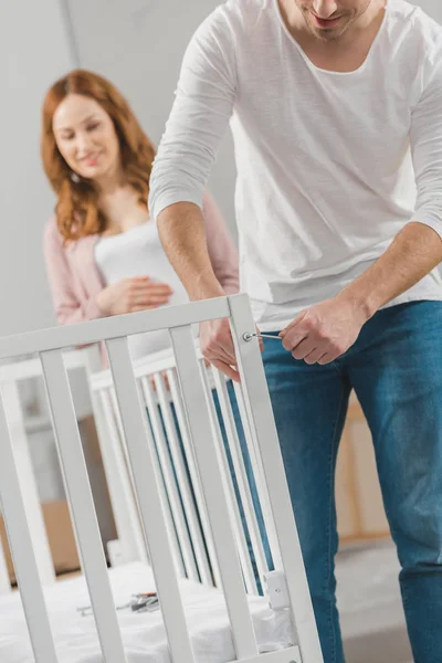 Cropped shot of man fixing baby bed while pregnant woman standing behind — Stock Photo