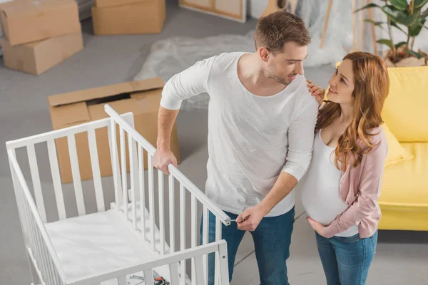 Vue grand angle de heureux jeune couple enceinte se souriant tout en se tenant près du lit de bébé — Photo de stock