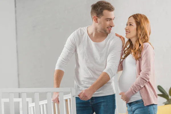 Glückliches junges schwangeres Paar lächelt einander an, während es neben dem Babybett steht — Stockfoto