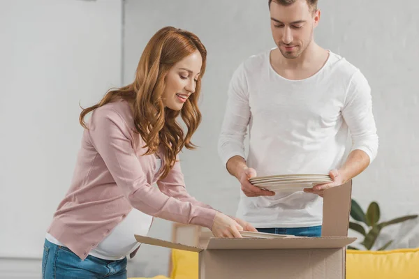 Young pregnant couple packing plates in cardboard box during relocation — Stock Photo