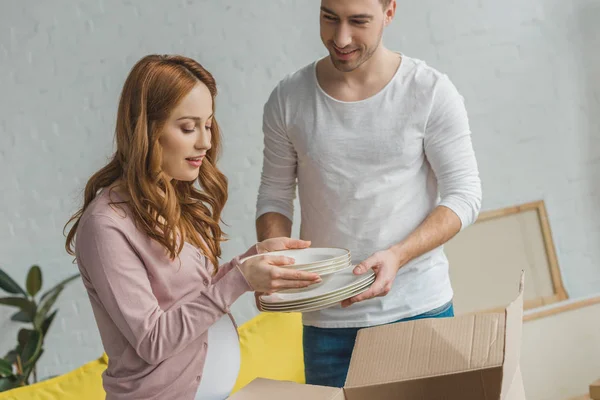 Junges schwangeres Paar packt Teller während des Umzugs in Karton — Stockfoto