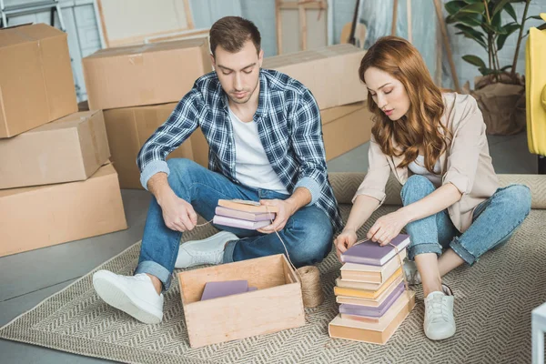 Jovem casal sentado no tapete e livros de embalagem na caixa durante a realocação — Fotografia de Stock