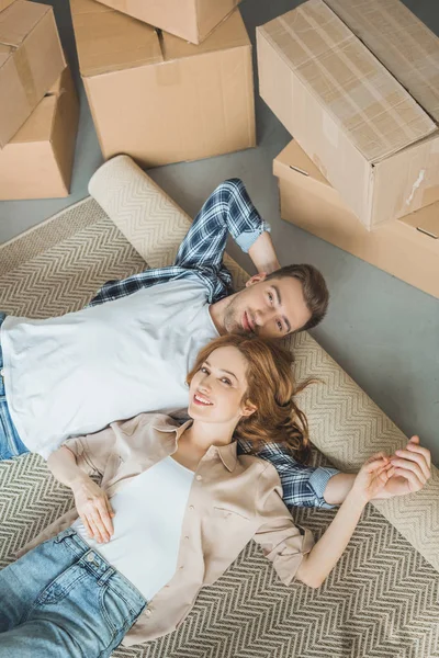 Vista superior de la joven pareja sonriendo a la cámara mientras está acostada en una alfombra enrollada cerca de cajas de cartón - foto de stock