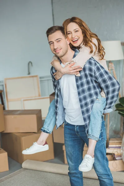 Smiling couple piggybacking at new apartment with cardboard boxes, relocation concept — Stock Photo