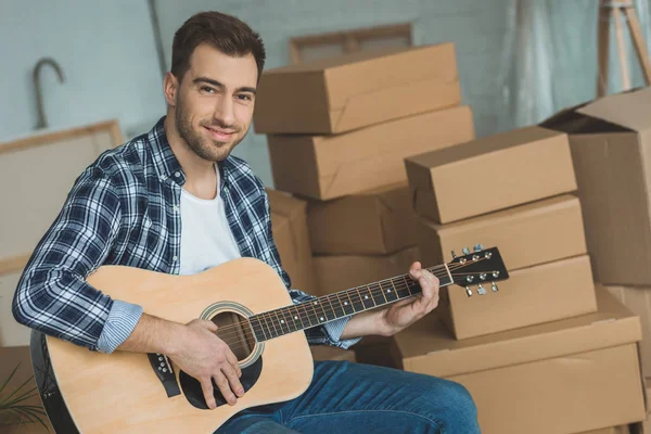 Portrait of smiling man with acoustic guitar at new home with cardboard boxes, relocation concept — Stock Photo