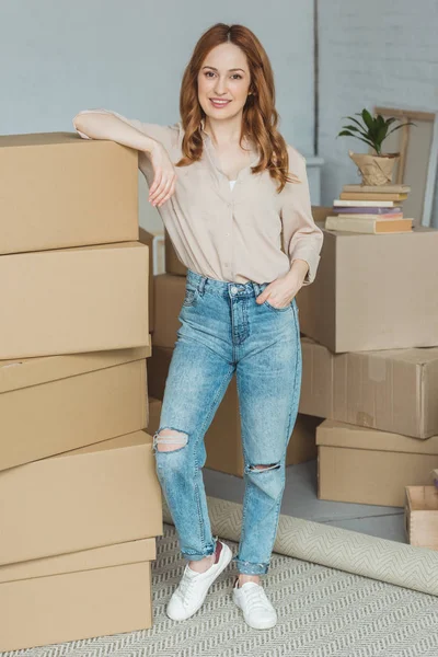 Femme souriante appuyée sur un tas de boîtes en carton à la nouvelle maison, concept de déménagement — Photo de stock