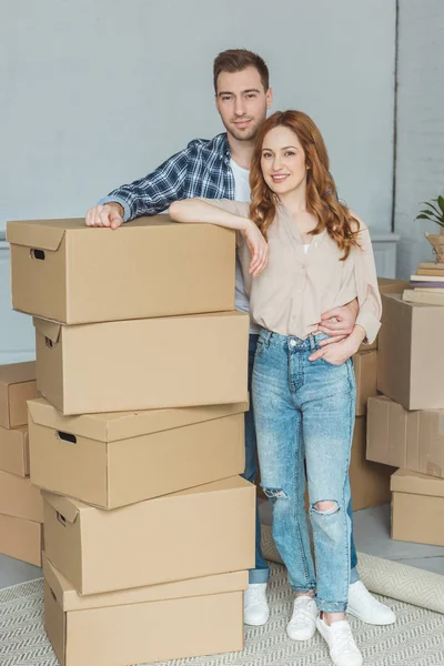 Sonriente pareja apoyada en la pila de cajas de cartón en el nuevo hogar, concepto de reubicación - foto de stock