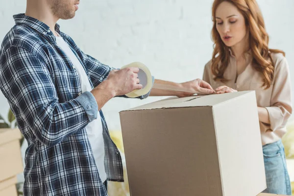 Partial view of couple packing cardboard box with sticky tape together, relocation concept — Stock Photo