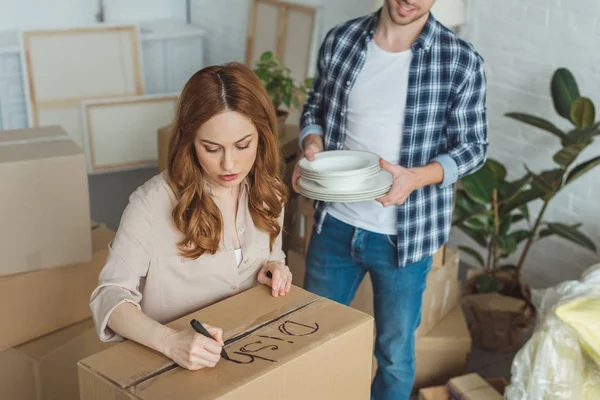 Частковий вид жінки, що підписує картонну коробку з чоловіком з посудом поруч, переміщуючи концепцію будинку — стокове фото