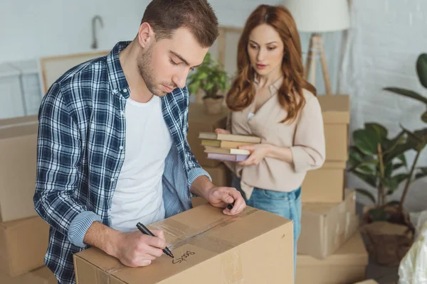 Man signing cardboard box with wife with books in hands near by, moving home concept — Stock Photo