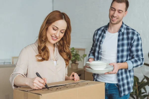 Femme souriante signant boîte en carton avec mari avec vaisselle à proximité, déménagement concept maison — Photo de stock