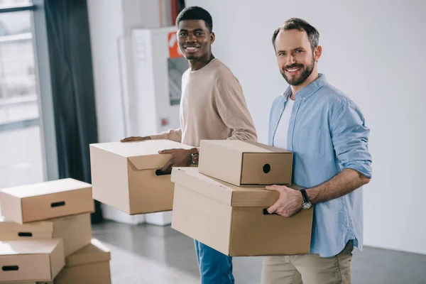 Compañeros de trabajo multiétnicos sosteniendo cajas de cartón y sonriendo a la cámara durante la reubicación - foto de stock