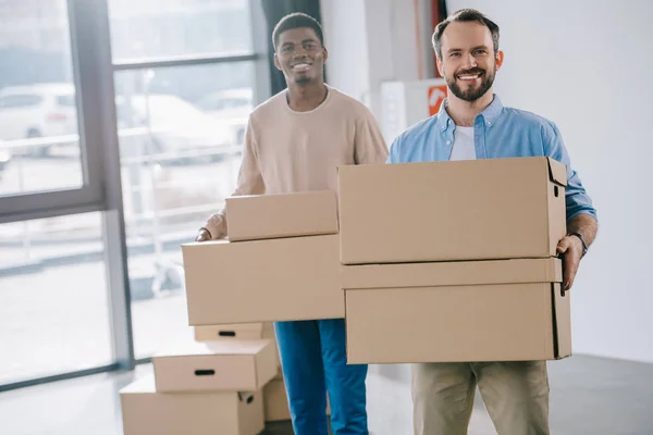 Glückliche multiethnische Männer, die Pappkartons in der Hand halten und beim Umzug in die Kamera lächeln — Stockfoto