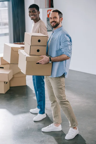 Jóvenes hombres multiétnicos sosteniendo cajas de cartón y sonriendo a la cámara durante la reubicación - foto de stock