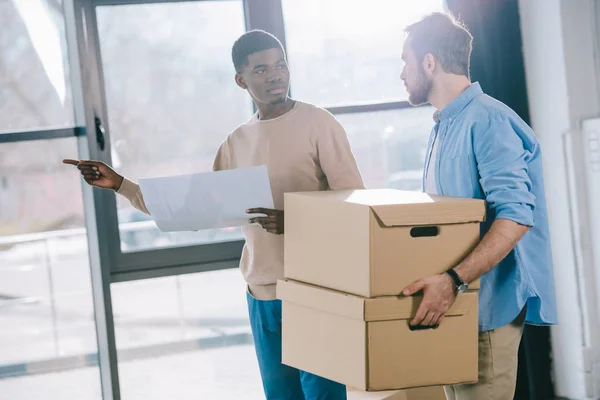 Jeunes hommes multiethniques qui se regardent tout en tenant du papier et des boîtes en carton pendant leur déménagement — Photo de stock